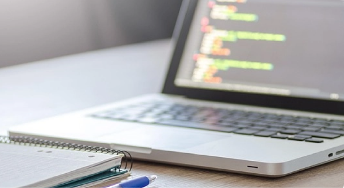 ノートパソコンの画面にソースコードが表示され、横にはリングノートとペンが置かれている。開発作業中のデスクの様子から、GISソフトウェアの構築やカスタマイズ支援サービスをイメージさせる構成。