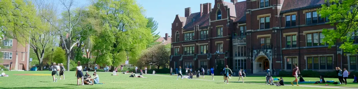 大学キャンパスの芝生広場で学生が交流や読書をしている様子。赤レンガの校舎と緑豊かな環境が、教育・学習の場としての空間を表現している。